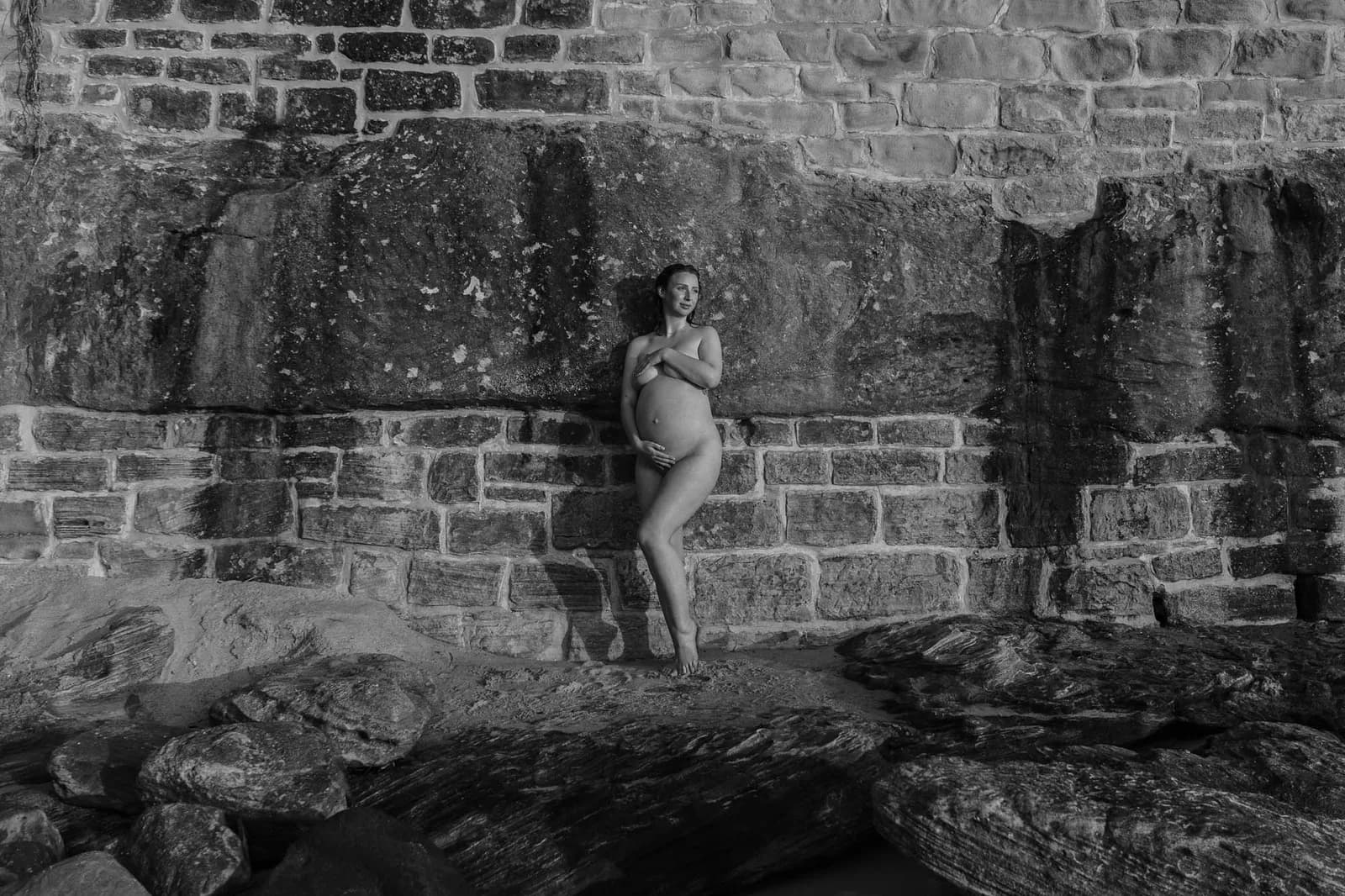 Wide black and white maternity portrait against a sandstone sea wall on the Sydney coast