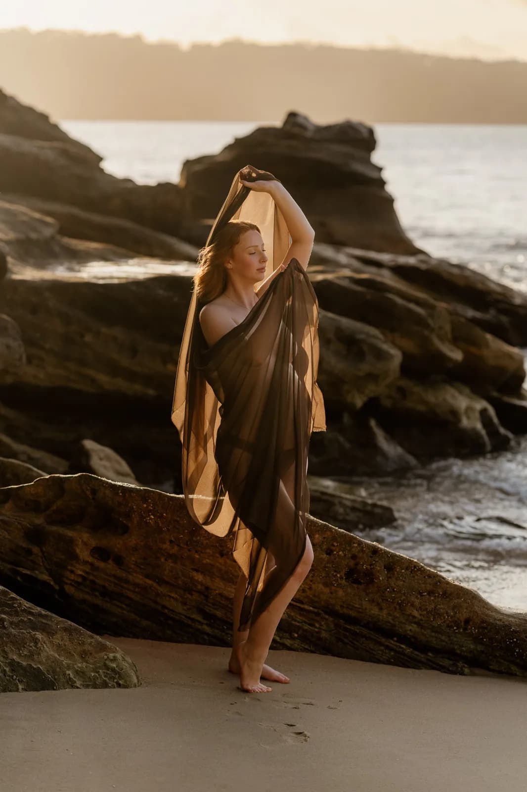 Pregnant woman wrapped in sheer fabric on the beach during a Sydney maternity photoshoot