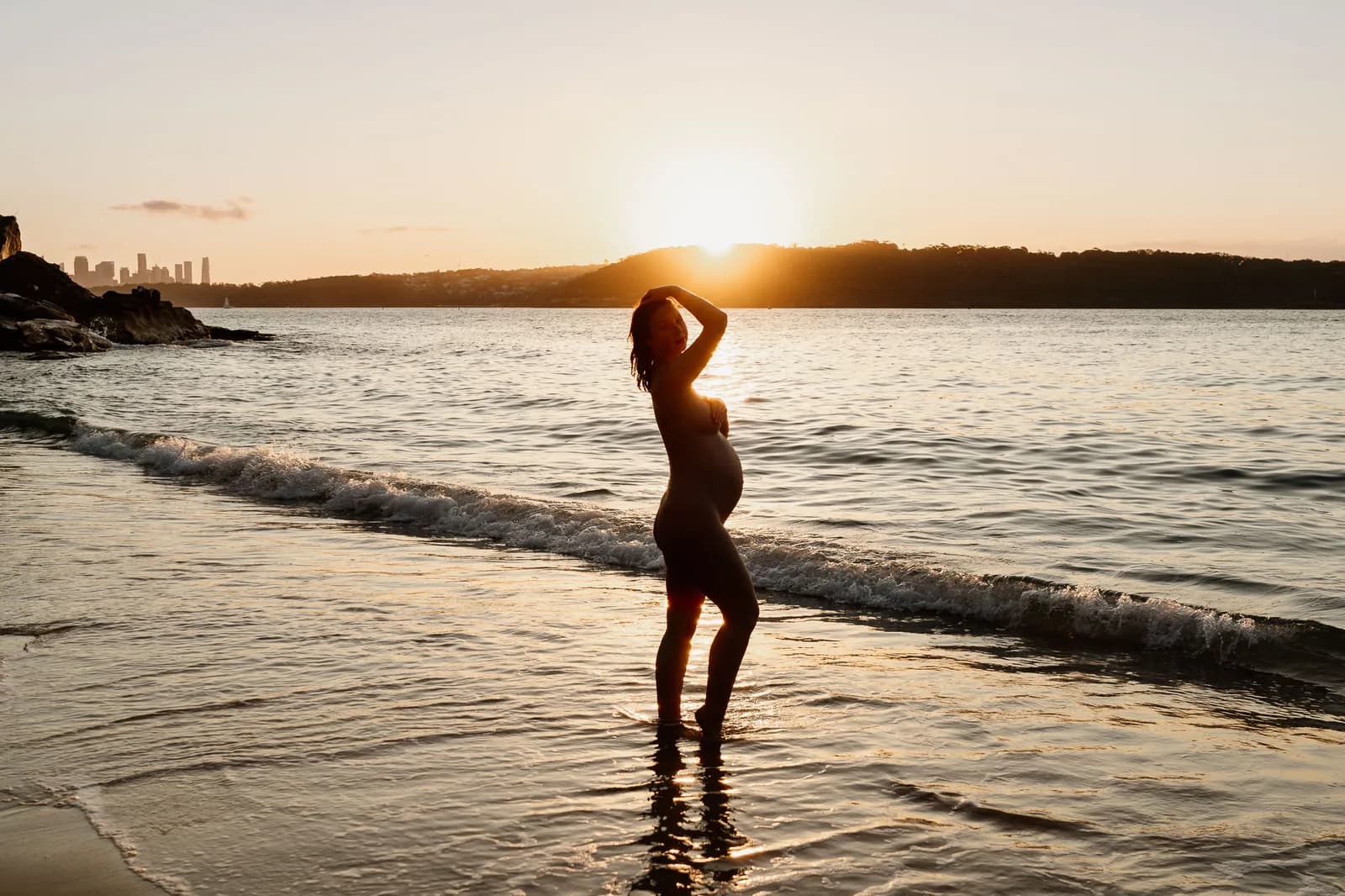 Expectant mother standing in the shallows at sunset during a nude maternity photoshoot on the Sydney coast
