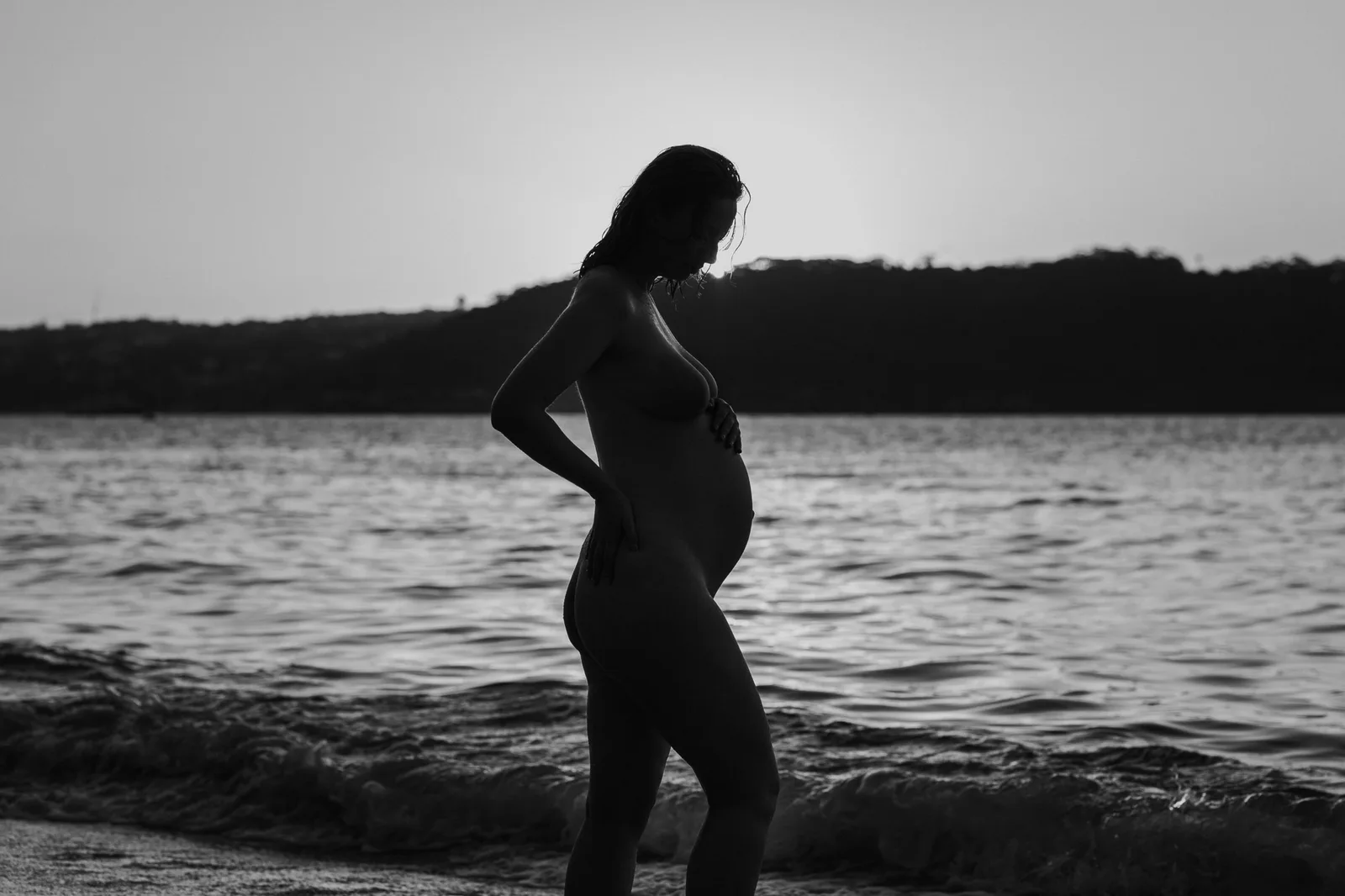Black and white silhouette of a pregnant woman standing in the water at dusk
