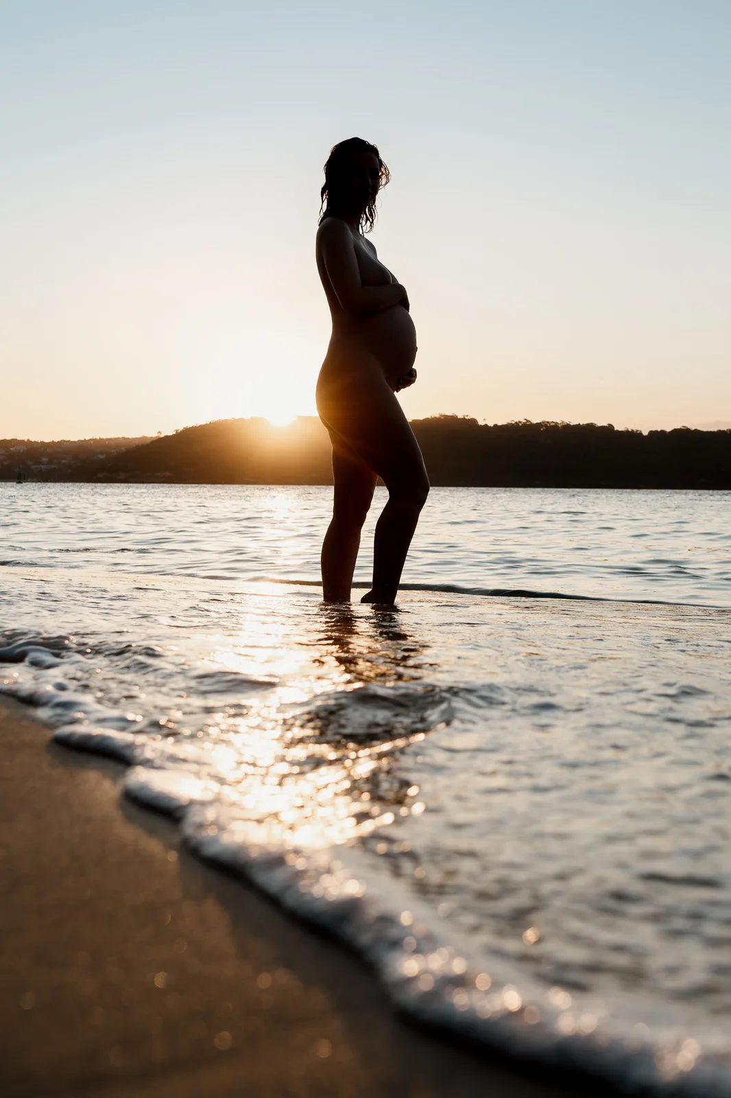 Silhouette of an expectant mother standing in the shallows at sunset during a Sydney maternity photoshoot
