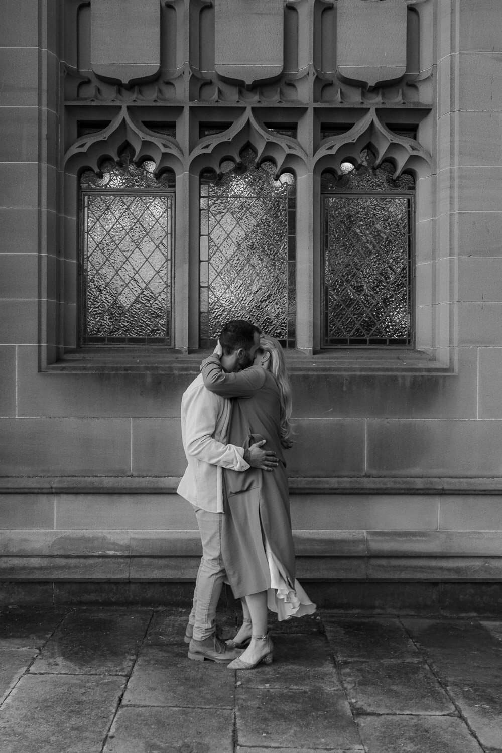 Black and white photograph of a couple embracing beside a sandstone church window