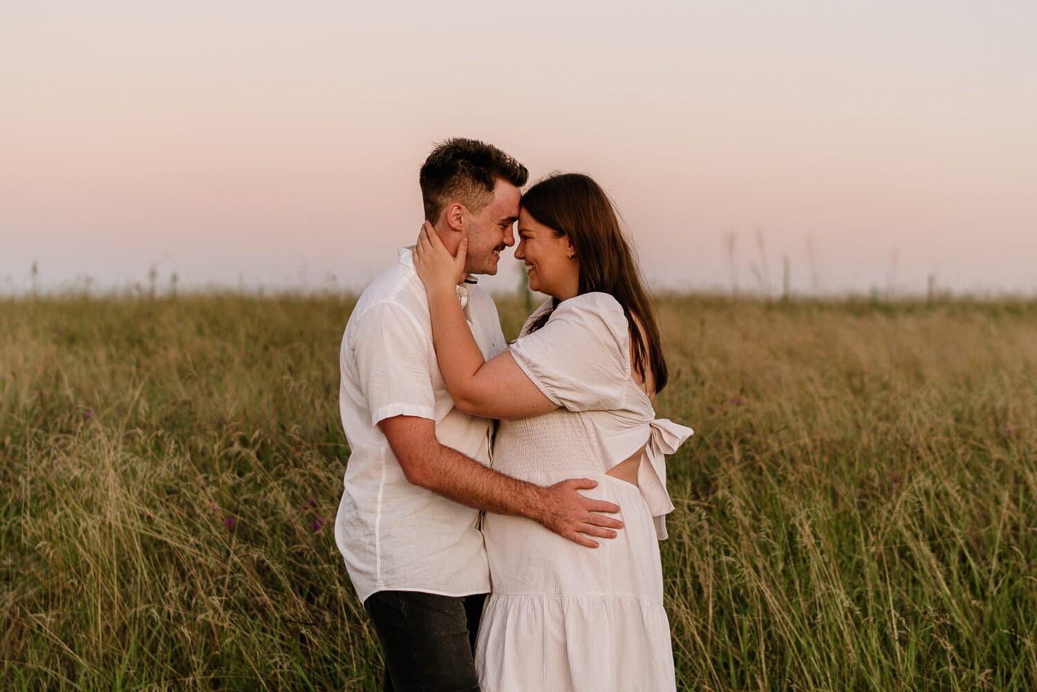 Couple embracing in tall grass under a soft pink sky