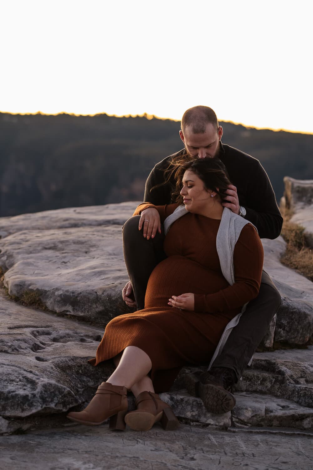 Expecting couple seated together on coastal rocks at sunset
