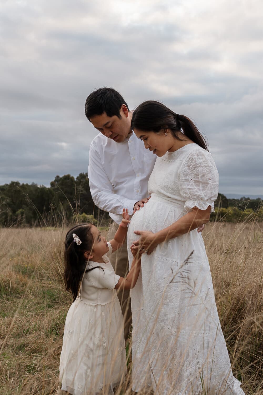 Expecting parents with their young daughter touching the baby bump in a golden field