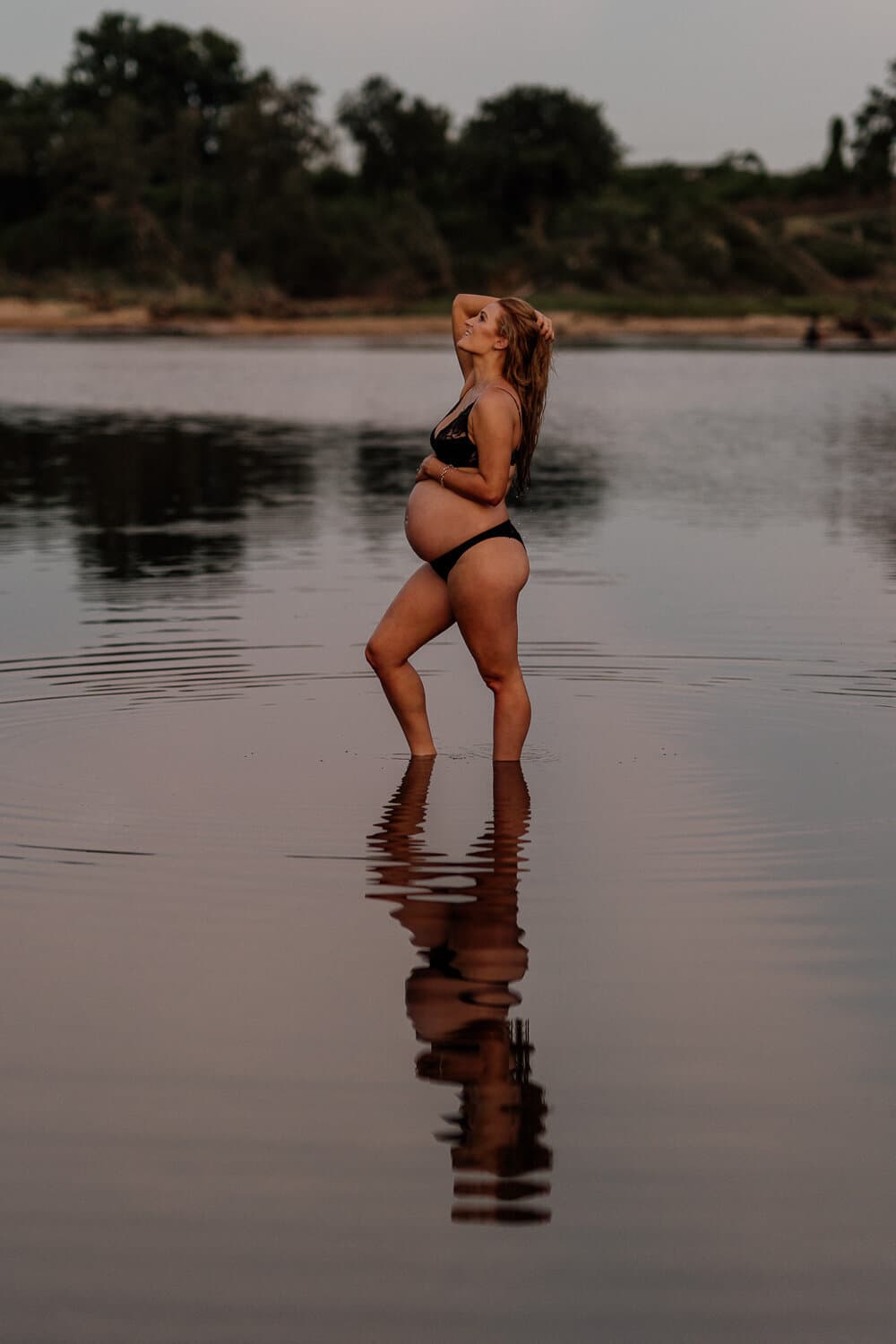 Mother-to-be standing in still water with her reflection at golden hour