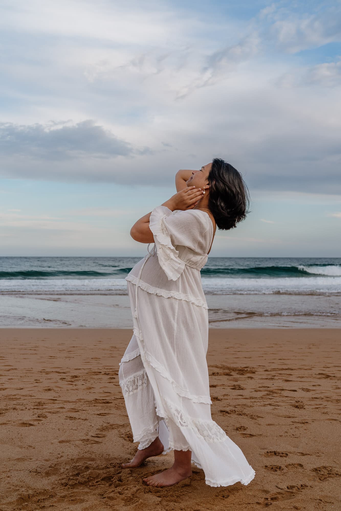 Mum-to-be in a flowing white dress on a Sydney beach at sunset