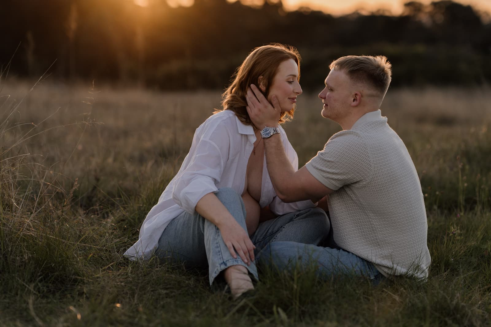 Expecting couple seated together in an open field, backlit by the setting sun