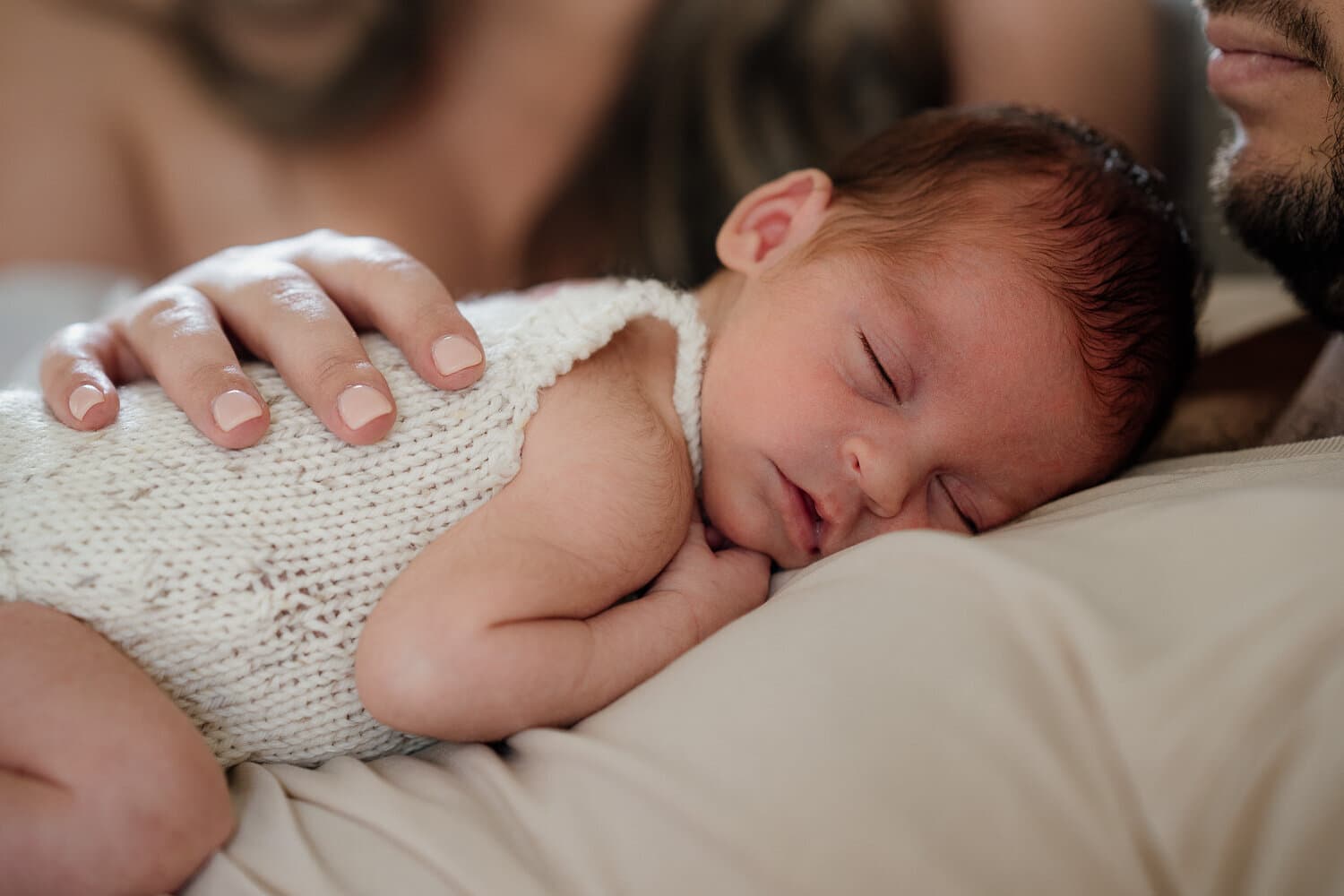 Sleeping newborn in a knitted romper with a parent's hand resting on their back