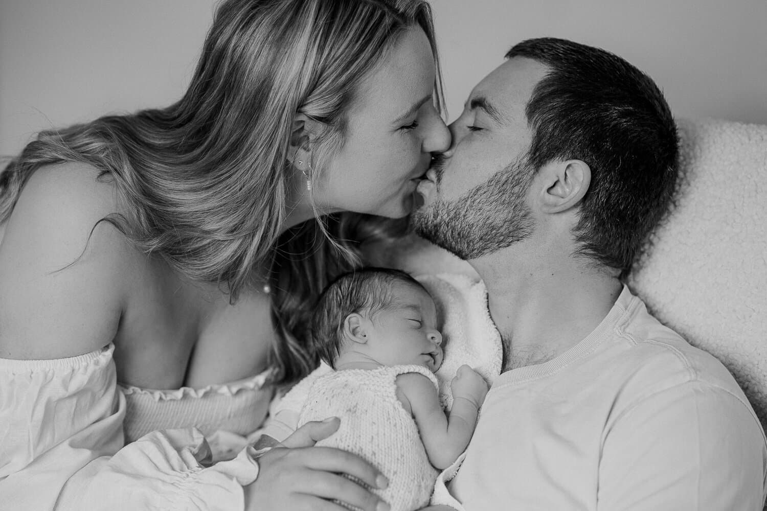 Black and white photograph of new parents kissing over their sleeping newborn