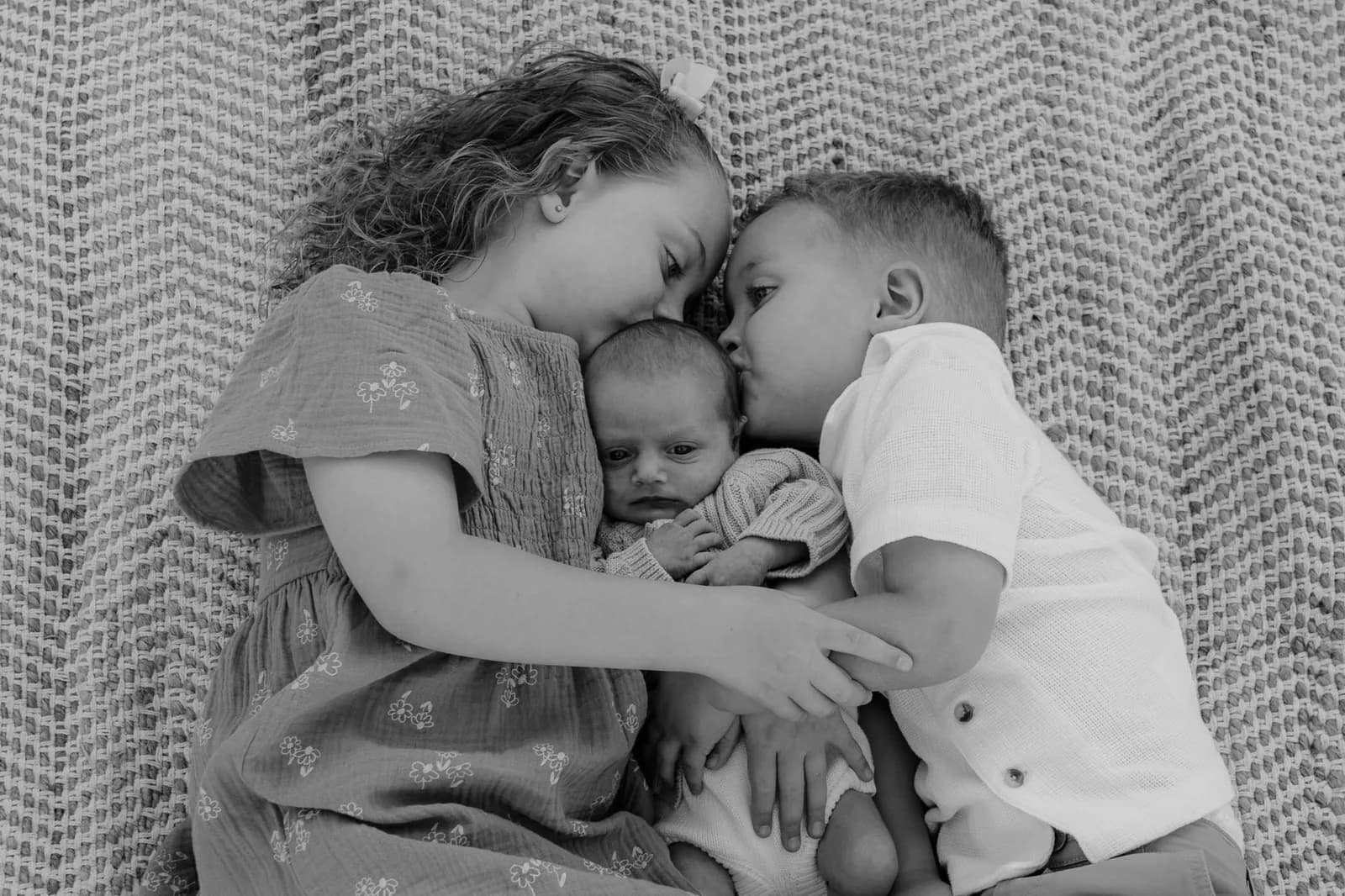 Black and white frame of 2 children leaning in to kiss their newborn sibling