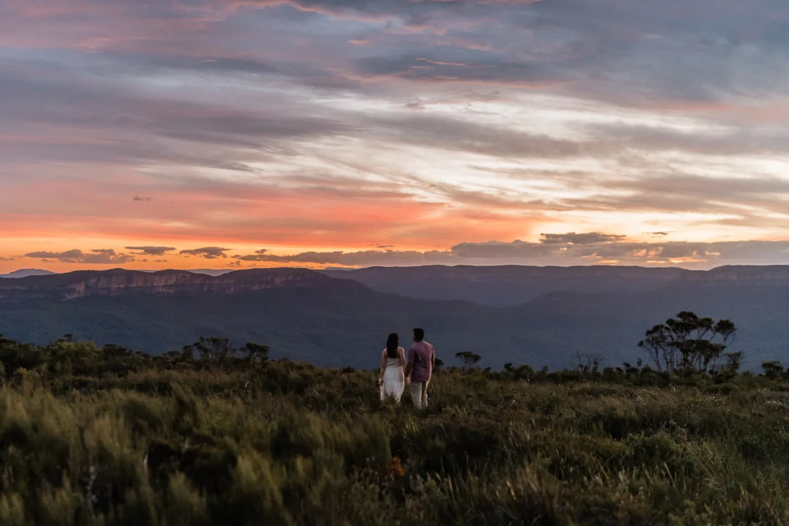 Couple standing small on a Blue Mountains clifftop as the sun sets behind the valley
