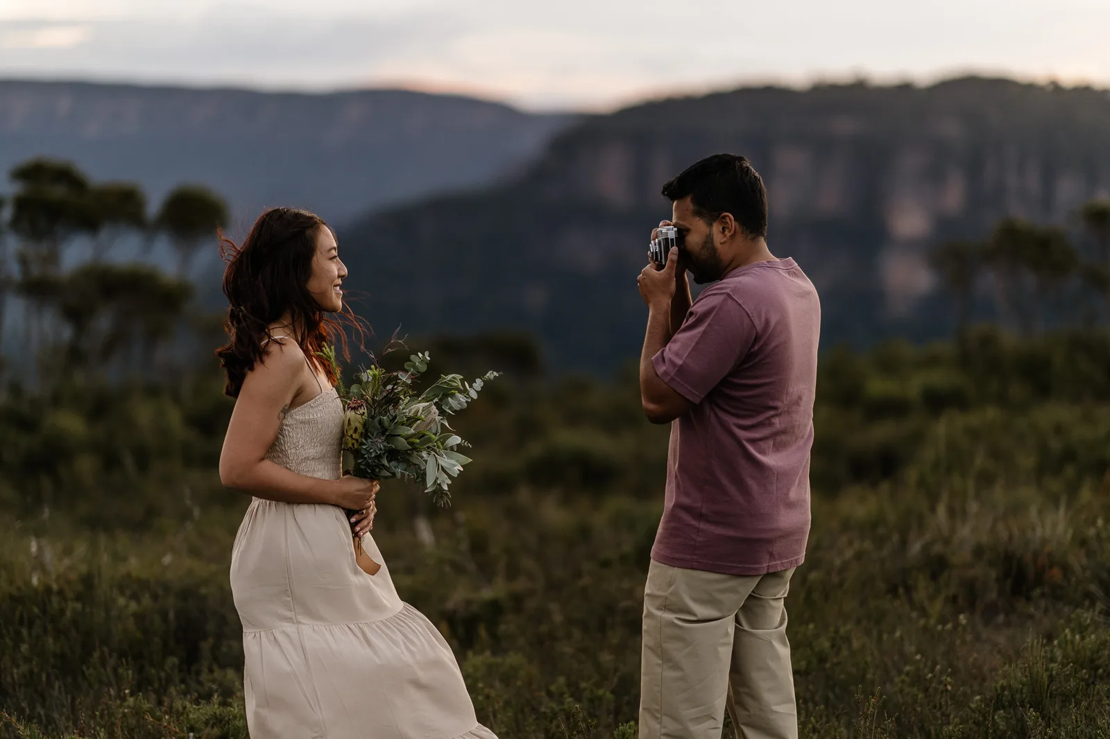 She holds flowers as he photographs her, with the Blue Mountains escarpment behind