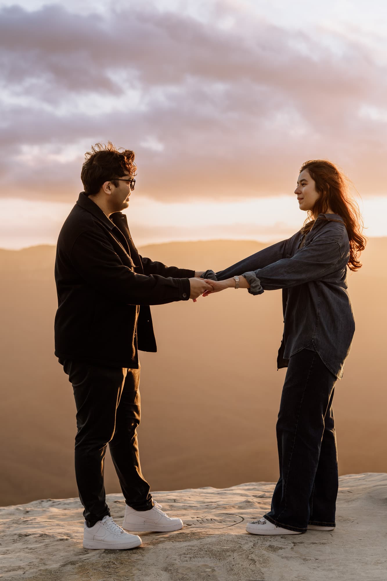 Newly engaged couple holding hands on a clifftop as the sun sets