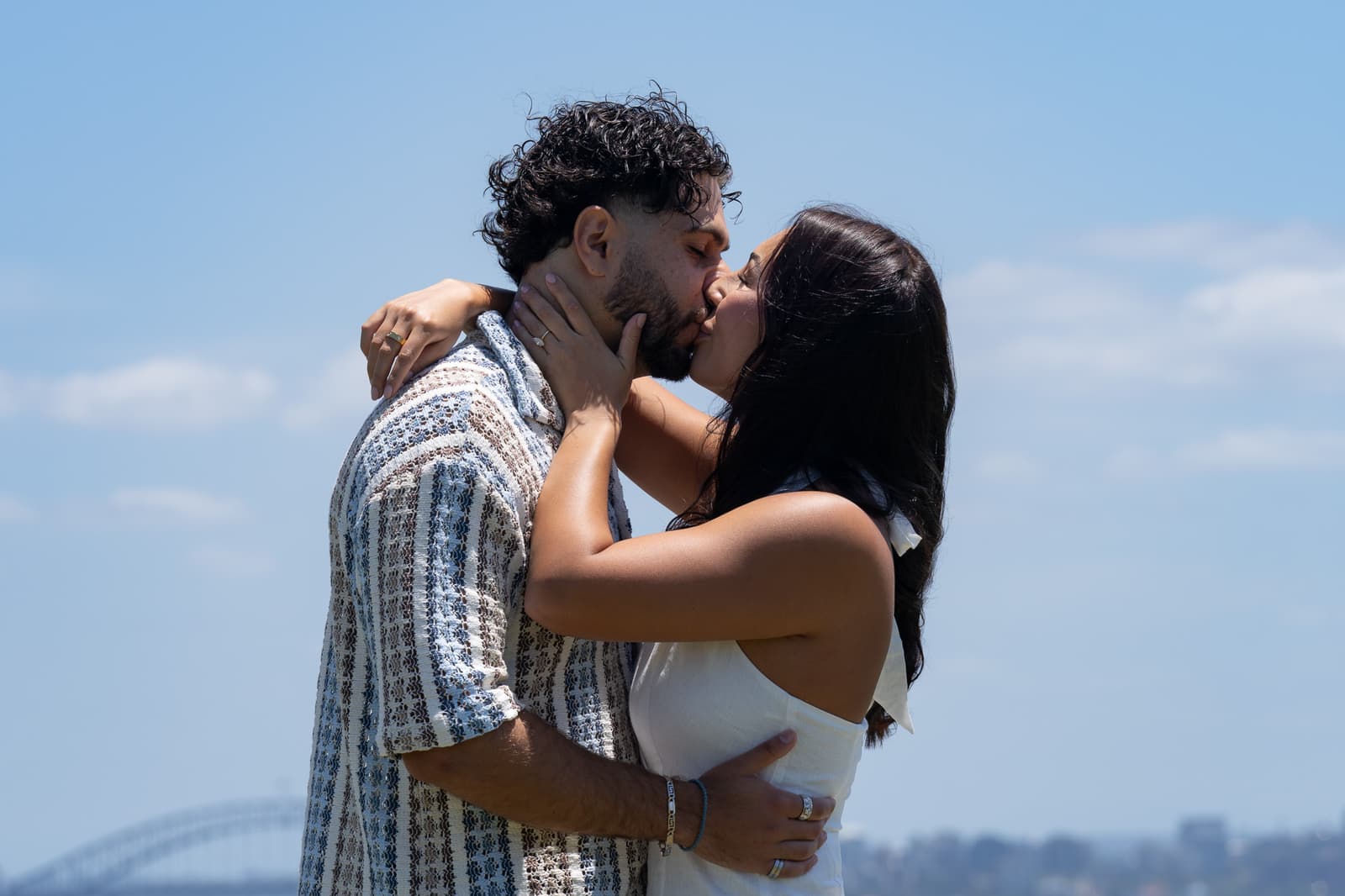 Newly engaged couple kissing under a clear blue sky