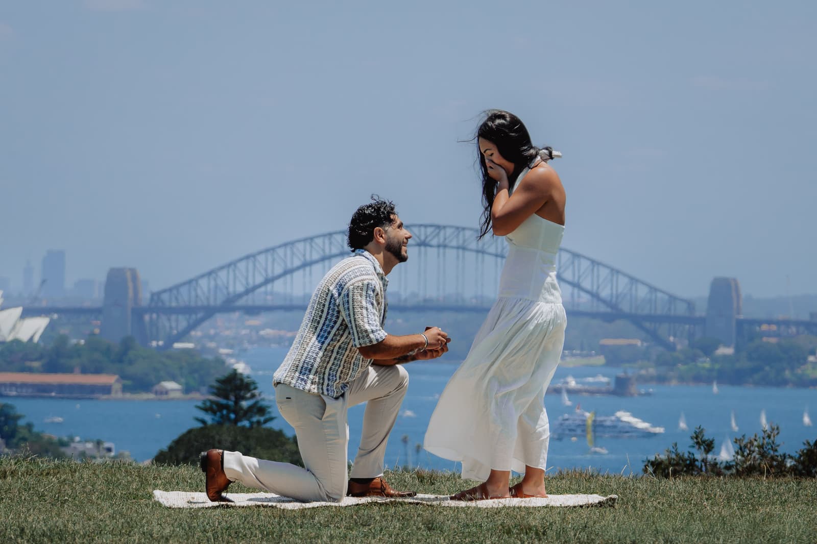 He kneels on the picnic rug to propose with the Harbour Bridge behind